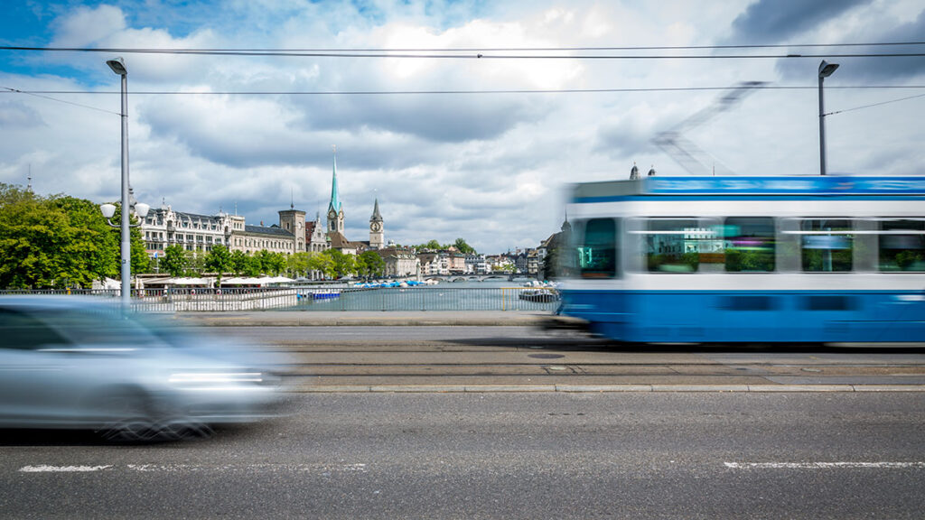 Blaues Tram auf der Quai-Brücke fährt vorbei und ein silbernes Auto kommt ihm entgegen. Im Hintergrund sieht man den Zürichsee.