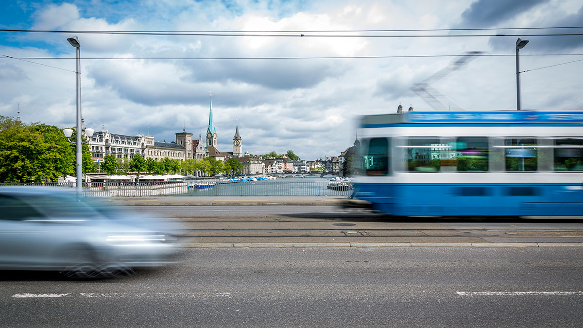 Blaues Tram auf der Quai-Brücke fährt vorbei und ein silbernes Auto kommt ihm entgegen. Im Hintergrund sieht man den Zürichsee.